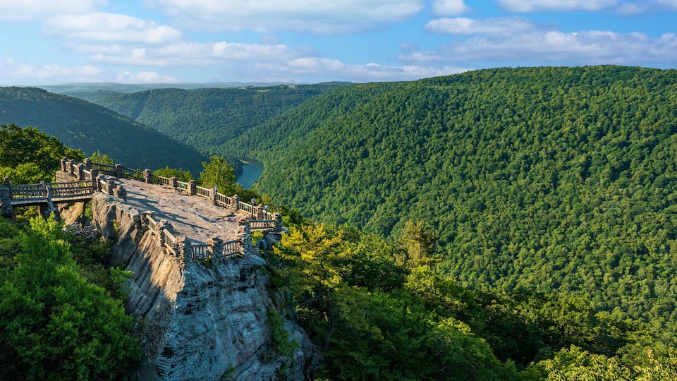 A stone bridge with a wooden railing overlooking a valley with dense forest and a river.