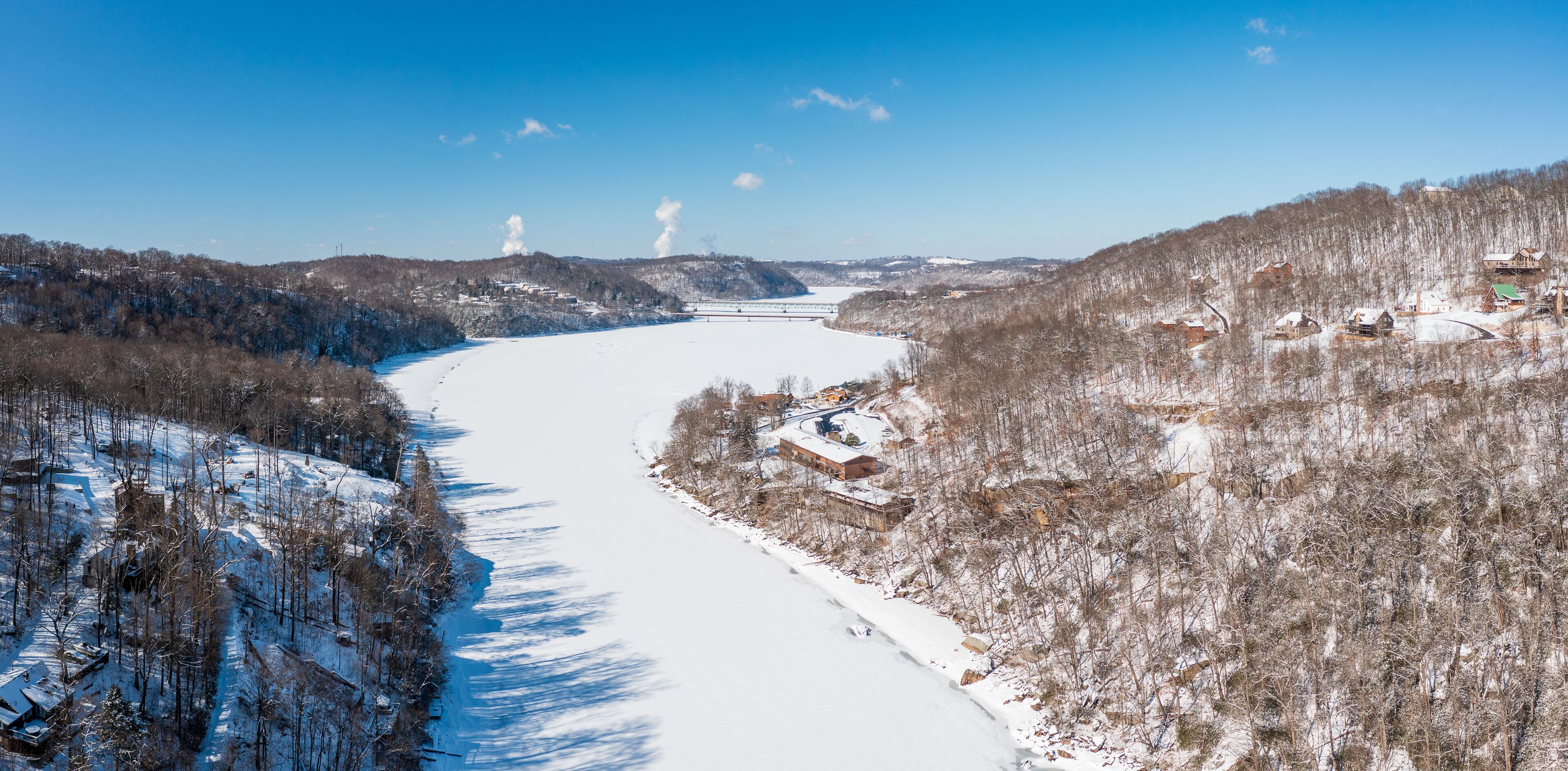 A snow-covered river valley with houses on the hillsides and a bridge in the distance.