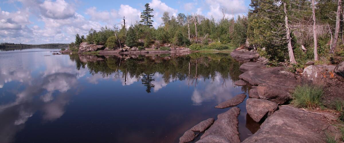 A calm lake with clear reflections of trees and sky, surrounded by rocky shores and dense forest.