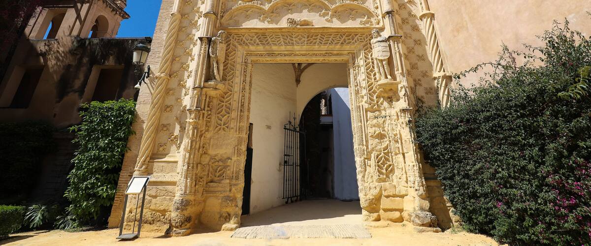 An ornate stone archway with intricate carvings and a barred window, leading into a building.