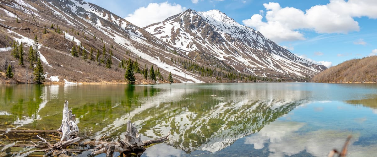 Panoramic view of St Elias Lake in northern Canada with calm, reflective water below snow capped peaks.