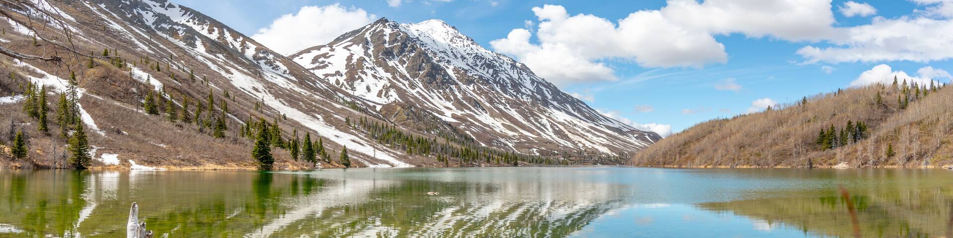 Panoramic view of St Elias Lake in northern Canada with calm, reflective water below snow capped peaks.