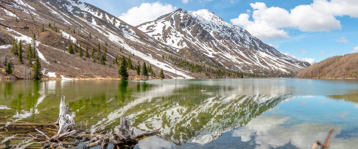 A mountain lake with clear reflections, surrounded by snow-capped peaks and coniferous trees.