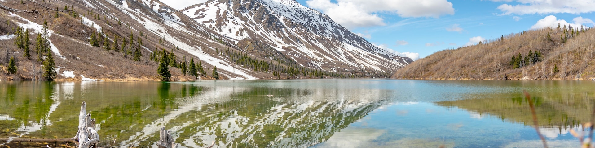 A mountain lake with clear reflections, surrounded by snow-capped peaks and coniferous trees.