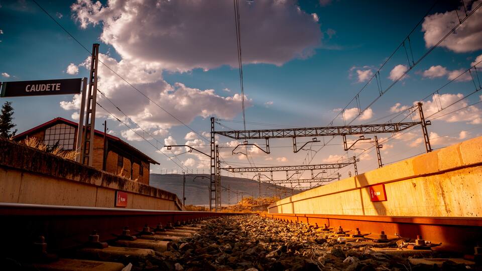 A railway station with overhead electric lines, a sign reading "CAUDETE," and a mountainous landscape in the background.