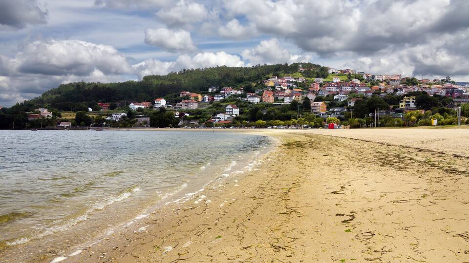 A beach with clear water, a sandy shore, and a hillside with colorful houses in the background.