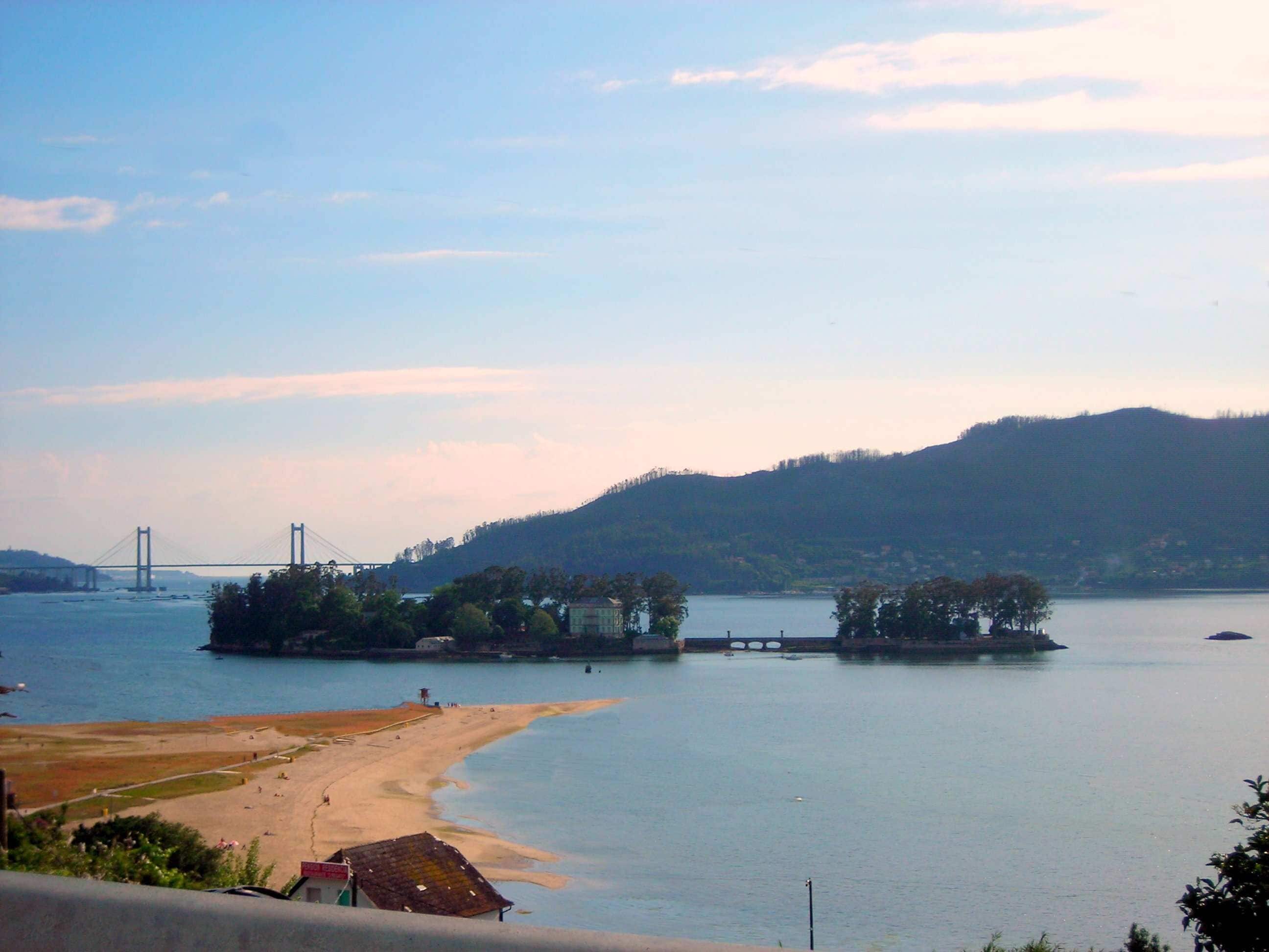 A coastal landscape with a beach, a bridge, and a forested hill.