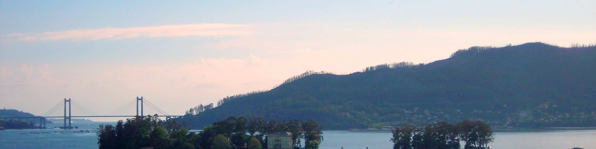 Un paisaje costero con playa, un puente y una colina boscosa.