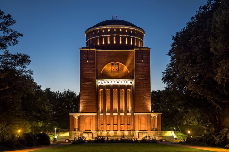 Un edifici de maó gran amb una cúpula i la paraula 'PLANETARIUM' a la façana, envoltat d'arbres i un camí ben il·luminat.