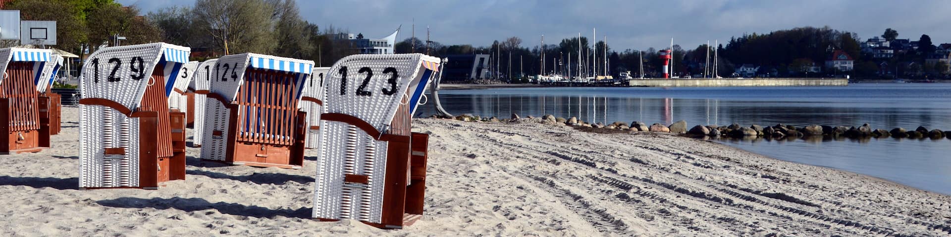 Beach chairs numbered 123, 124, and 129 on a sandy beach with a calm body of water and a cloudy sky in the background.
