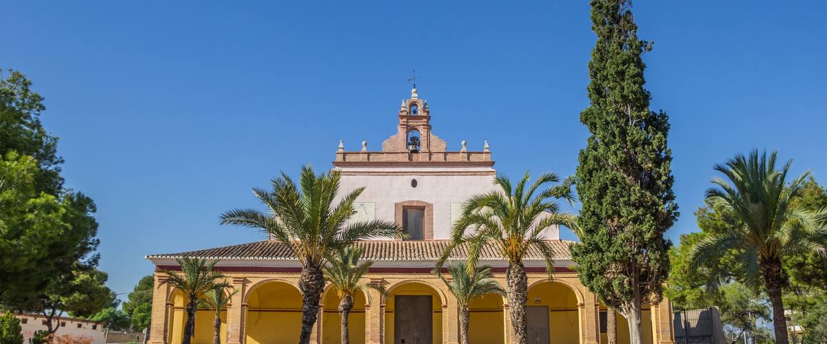 A historic church with a bell tower, surrounded by palm trees and a clear blue sky.
