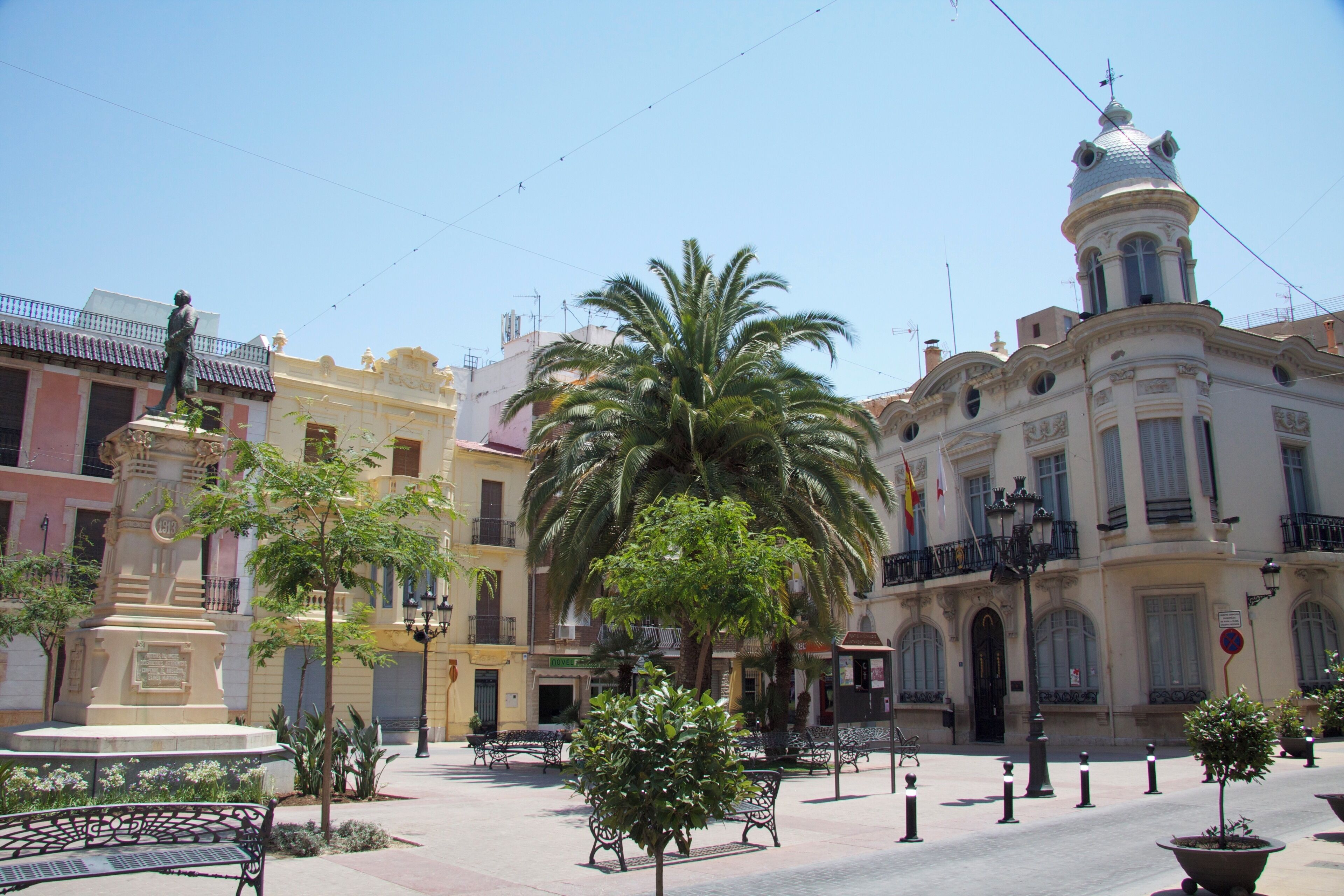 A plaza with a palm tree, benches, a statue, and historic buildings.