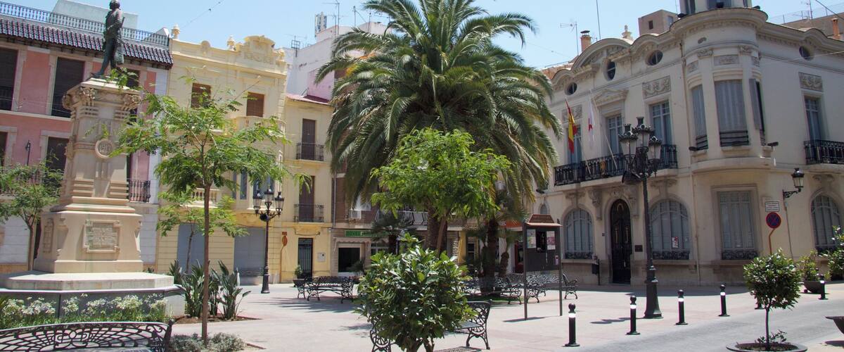 A plaza with a palm tree, benches, a statue, and historic buildings.