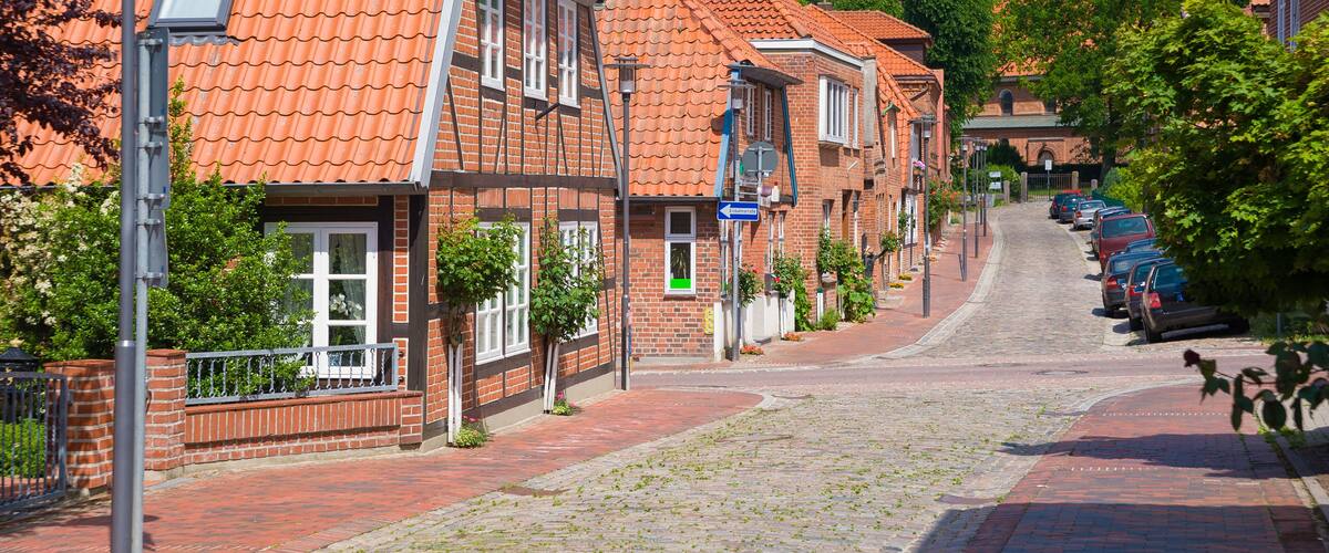 A cobblestone street lined with brick buildings, a church with a tower, and parked cars.