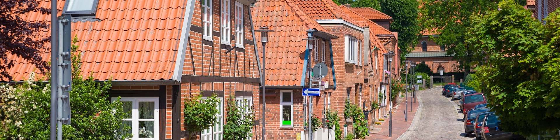 A cobblestone street lined with brick buildings, a church with a tower, and parked cars.