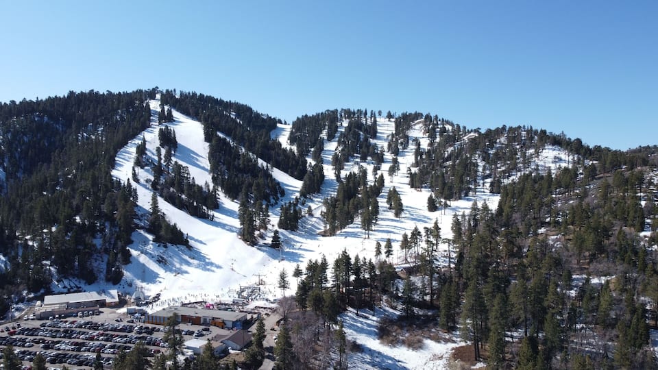 A snow-covered mountain with ski slopes, a parking lot, and a small building at the base.