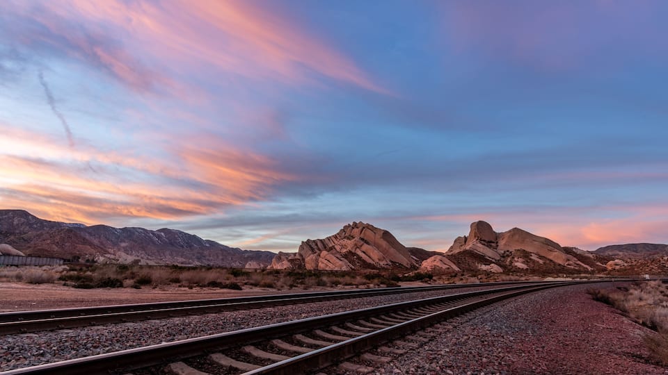A railway track leading through a desert landscape with rocky mountains in the background.