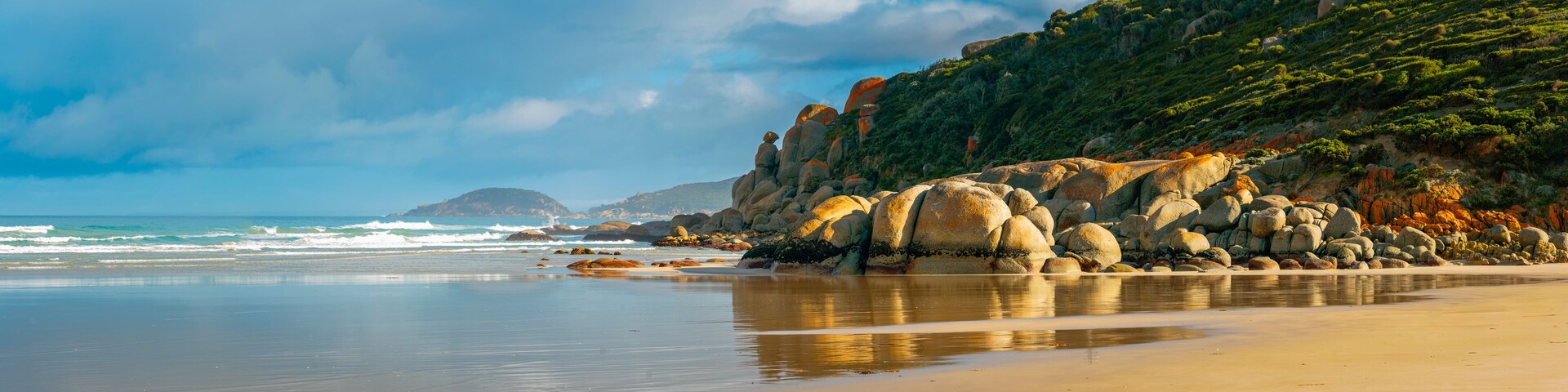 Una playa con grandes rocas, mar tranquilo y un promontorio cubierto de bosque.