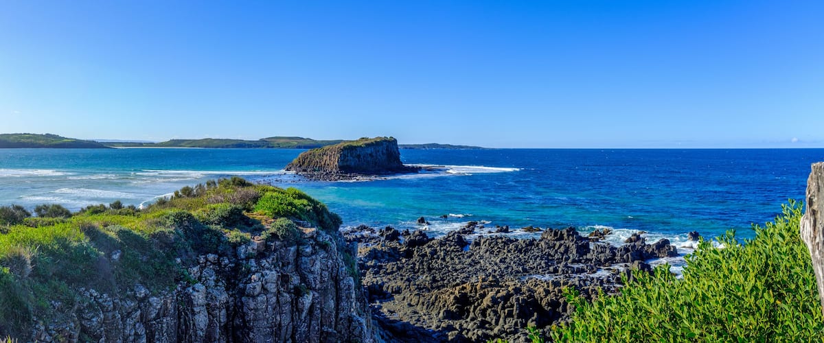 A coastal landscape with a prominent rocky outcrop, clear blue sea, and distant green hills.