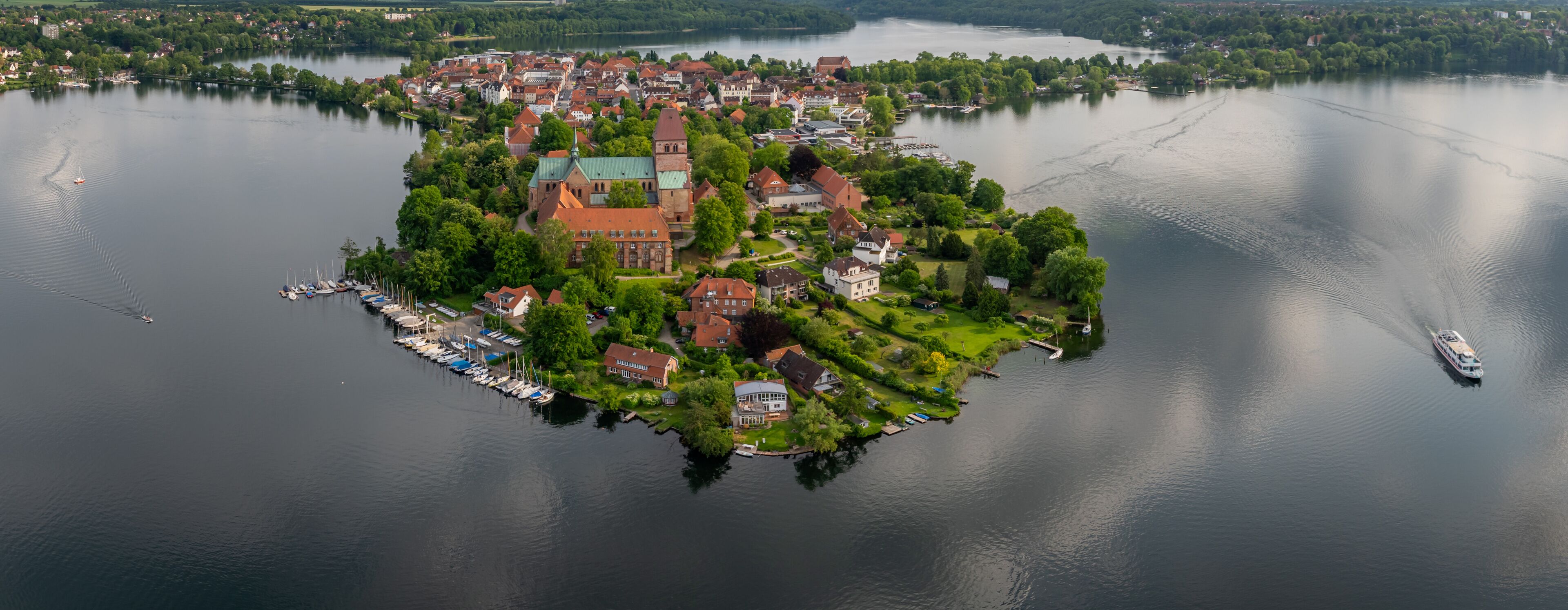 Ratzeburg’s old town island in northern Germany with cathedral and red-roofed houses, as the camera pans right to left showing the historic town, lake scenery and a sailing boat.