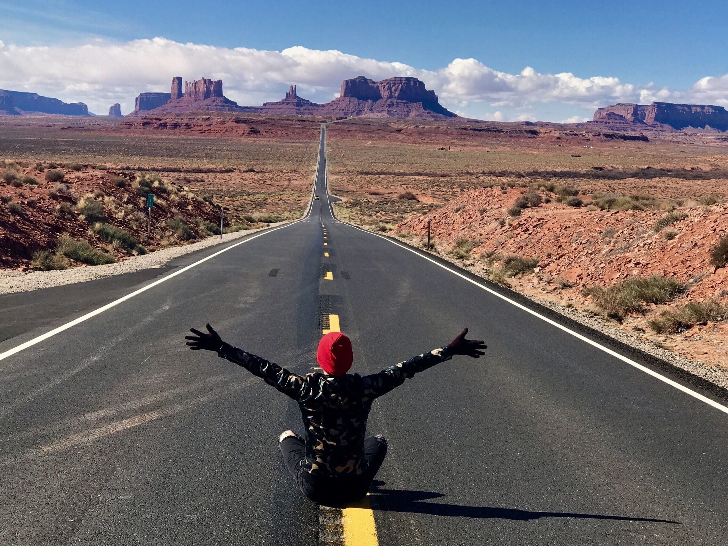 A long, straight road in a desert landscape with red rock formations in the distance.