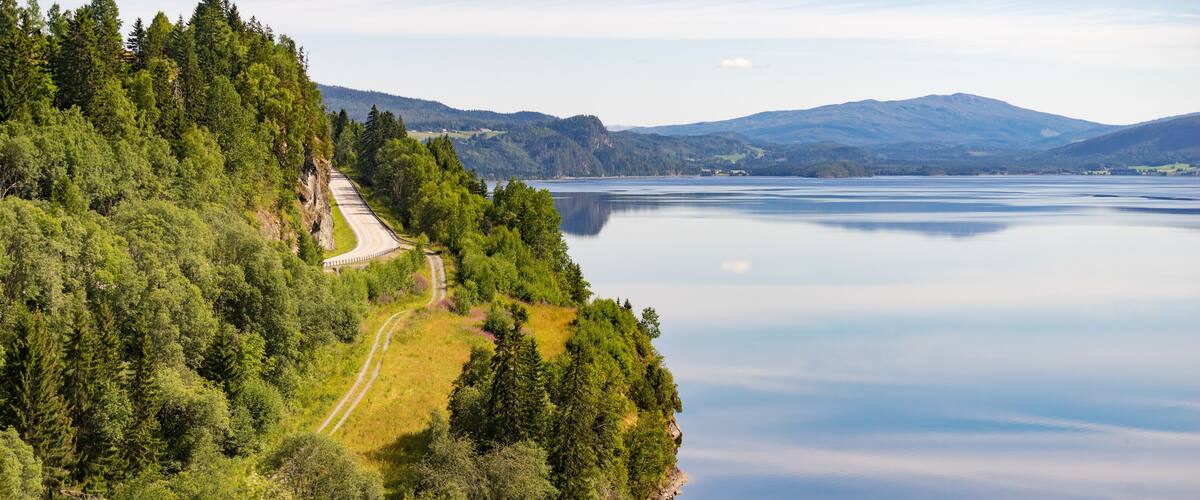 A winding road through a forested area with a lake and mountains in the background.