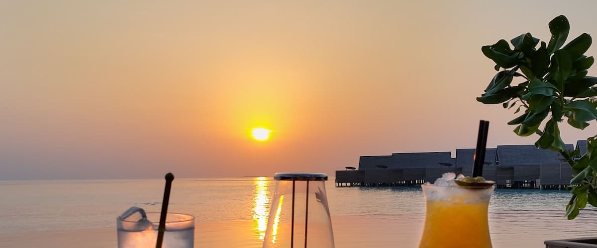 A sunset over a body of water with a wooden deck, two glasses of beverages, and a tropical drink.