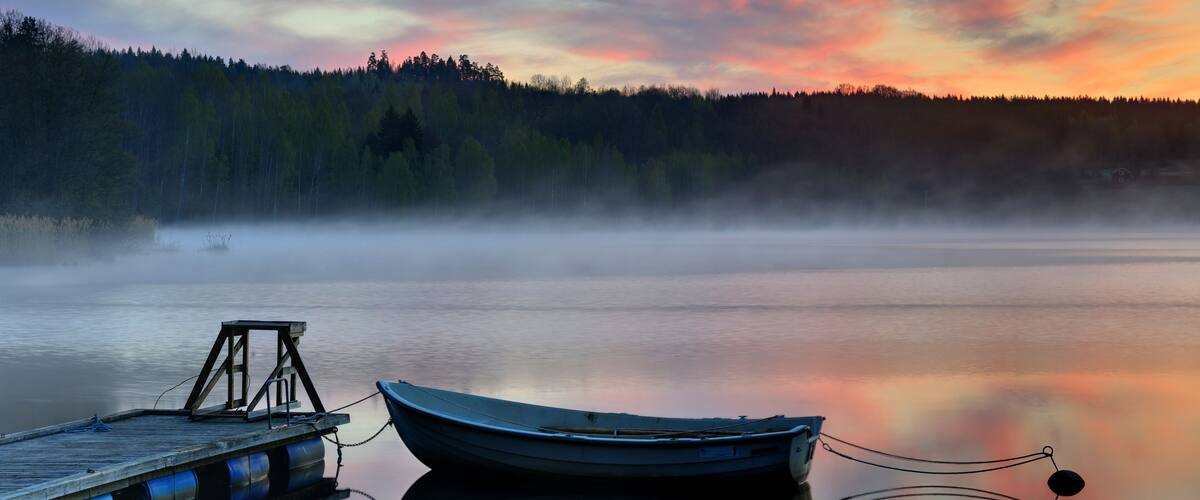 A calm lake at sunrise with a boat on a dock and a forest in the background.