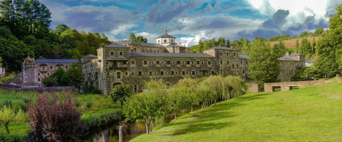 A historic stone building with a tower, surrounded by lush greenery and a river.