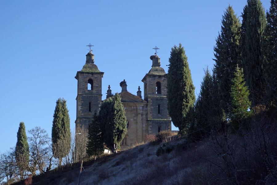Une église historique, dotée de deux tours de clocher et d’une façade en pierre, entourée d’arbres et située au pied d’une colline.