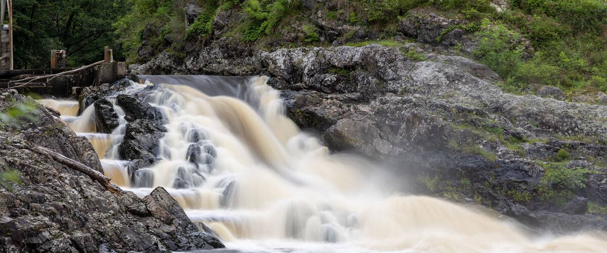 A waterfall cascading over rocks in a forested area with a bridge in the background.