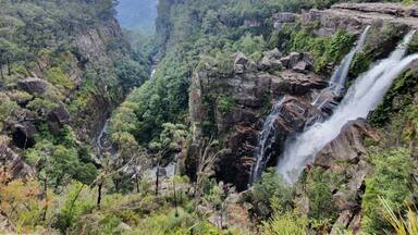 A lush valley with multiple waterfalls cascading down rocky cliffs, surrounded by dense greenery and forested hills.