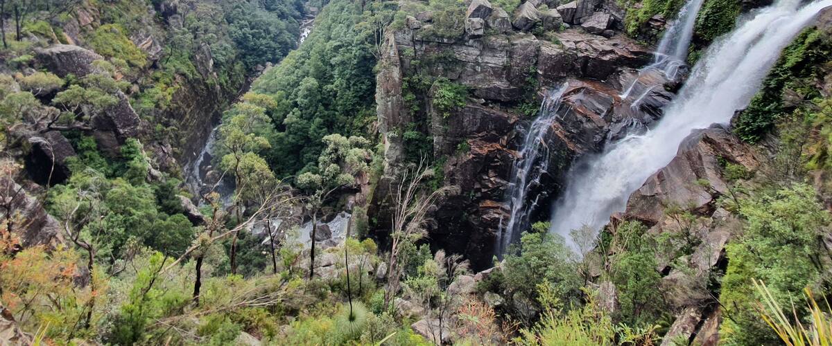 A lush valley with multiple waterfalls cascading down rocky cliffs, surrounded by dense greenery and forested hills.