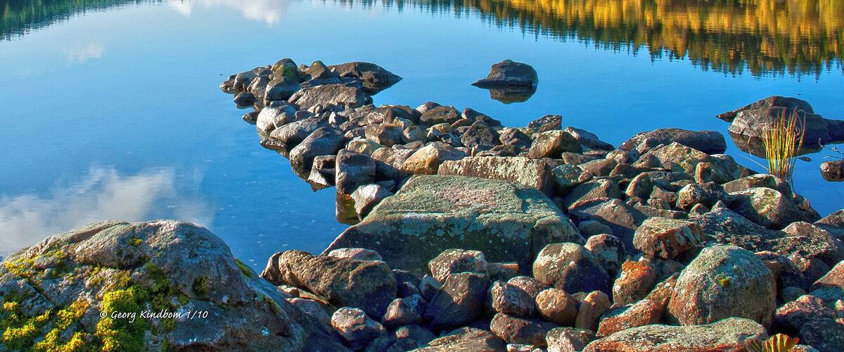 A calm lake with a rocky shoreline and a forested hill in the background.