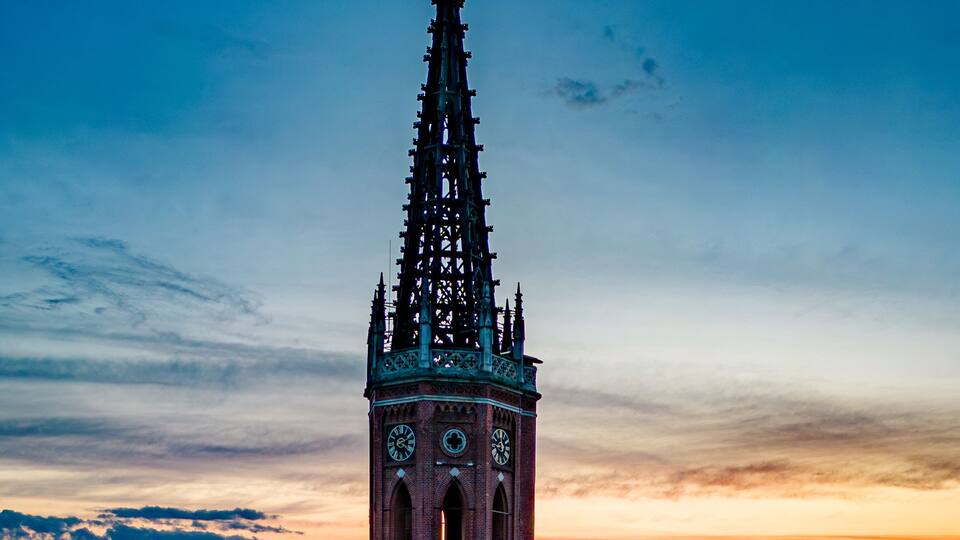 A tall brick tower with a spire, surrounded by greenery and buildings.