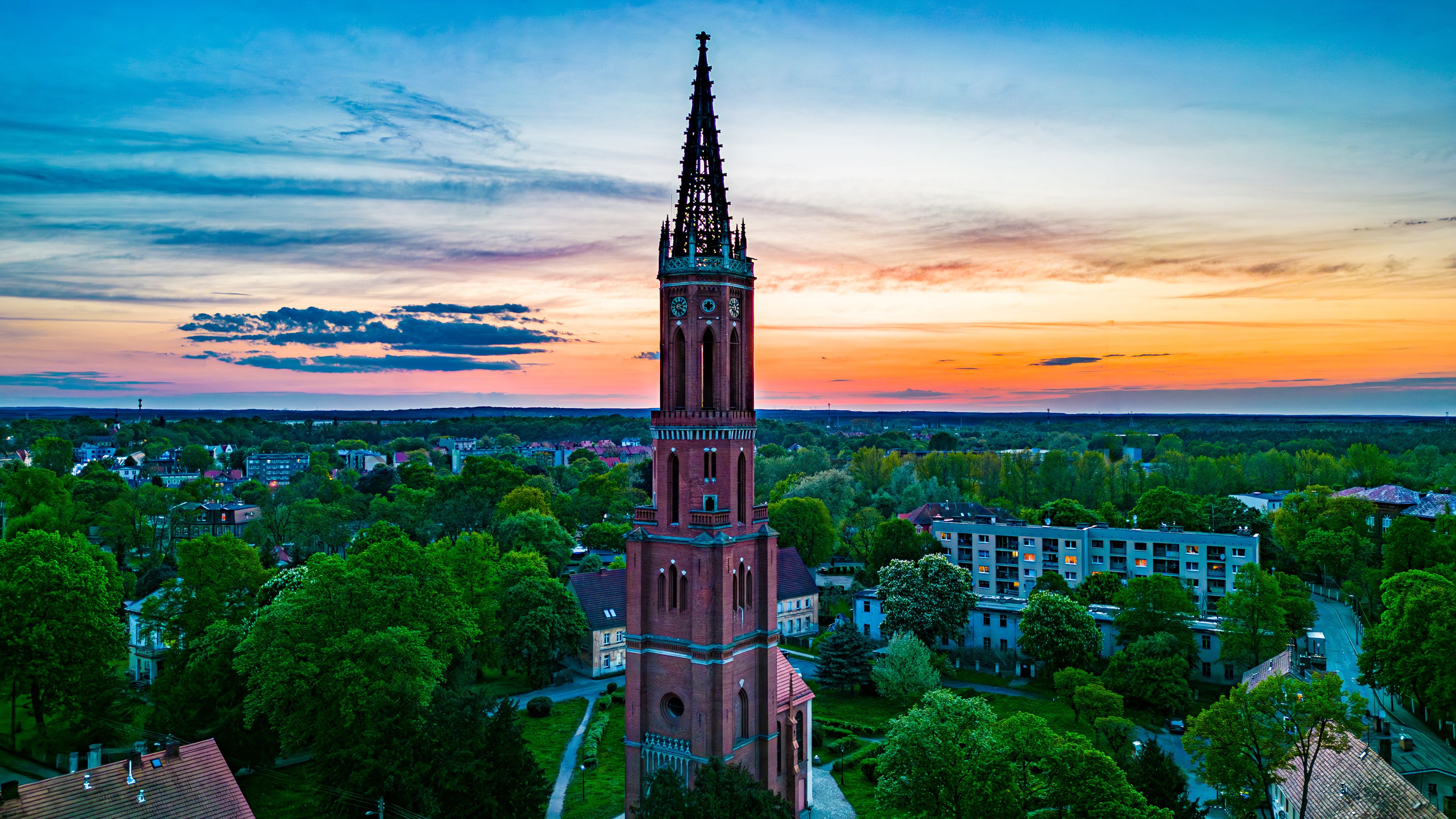 Majestic Church Tower at Sunset in Żagań, Poland. Aerial view of a grand church tower in Żagań, Poland, surrounded by lush greenery and urban scenery, set against a breathtaking sunset sky.