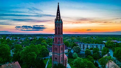 Majestic Church Tower at Sunset in Żagań, Poland. Aerial view of a grand church tower in Żagań, Poland, surrounded by lush greenery and urban scenery, set against a breathtaking sunset sky.