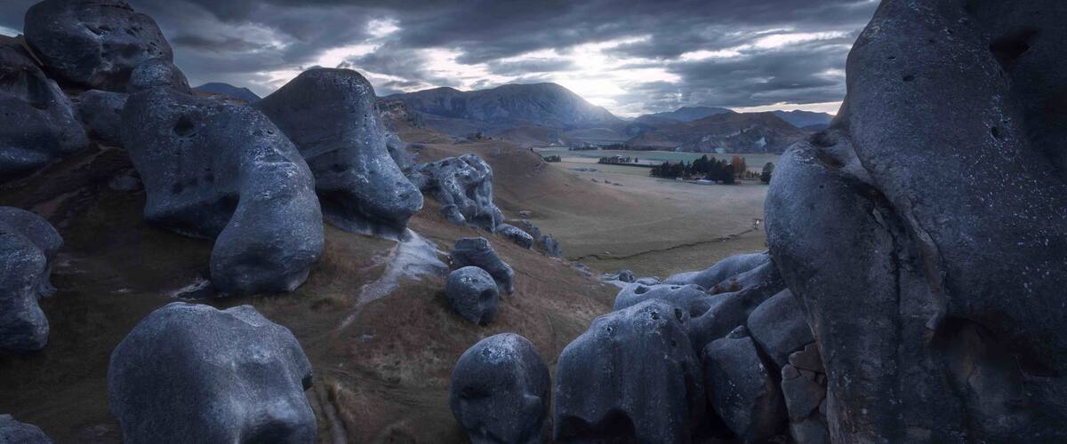 A rocky landscape with large boulders and a distant view of mountains under a cloudy sky.