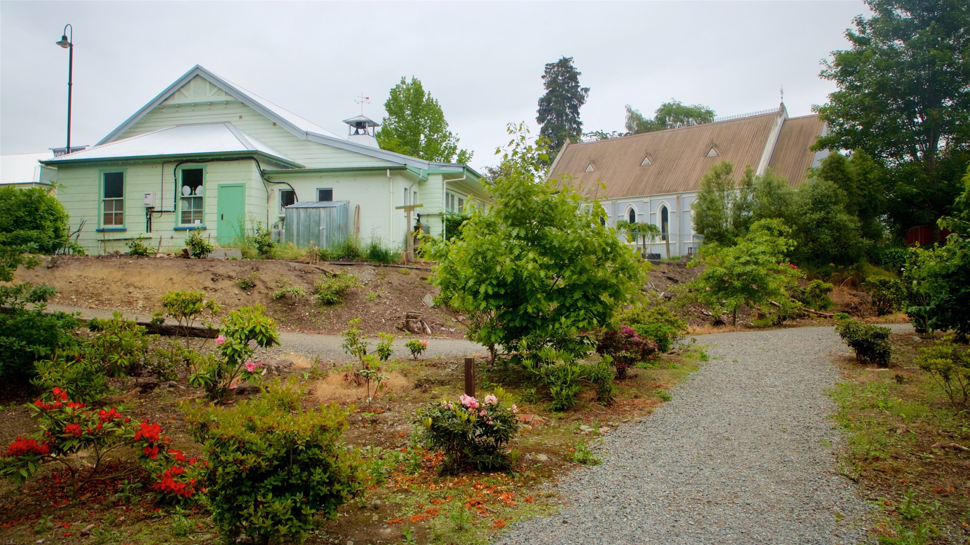 A rural landscape with a gravel path, green and white houses, and a variety of shrubs and flowers.