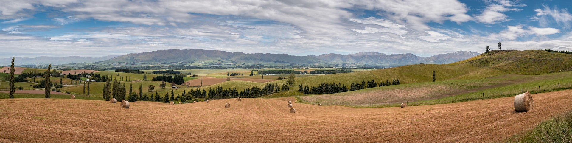 Panoramic view from the Fairlie - Geraldine lookout of straw bales sitting in a field in the rolling hills near Fairlie in the Canterbury region of New Zealand with Mount Dobson in the background
