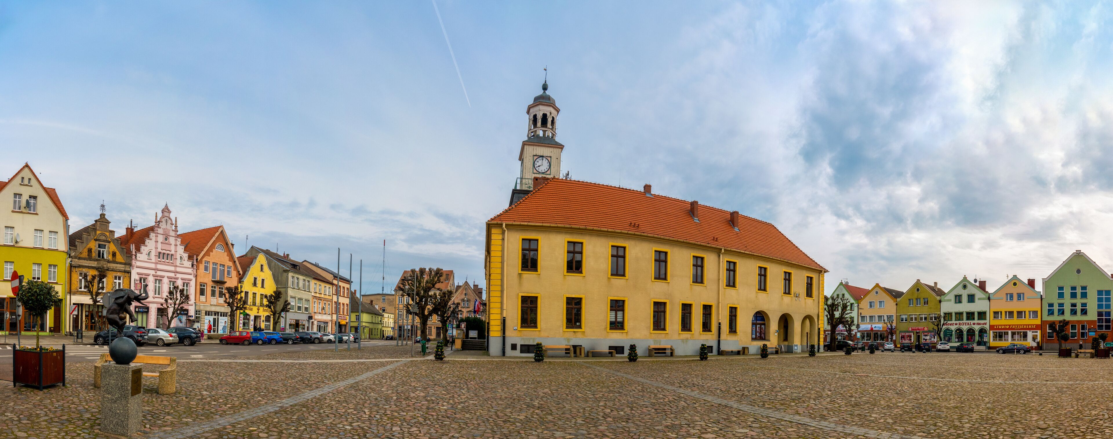 A cobblestone square with colorful buildings, a prominent clock tower, and a statue.
