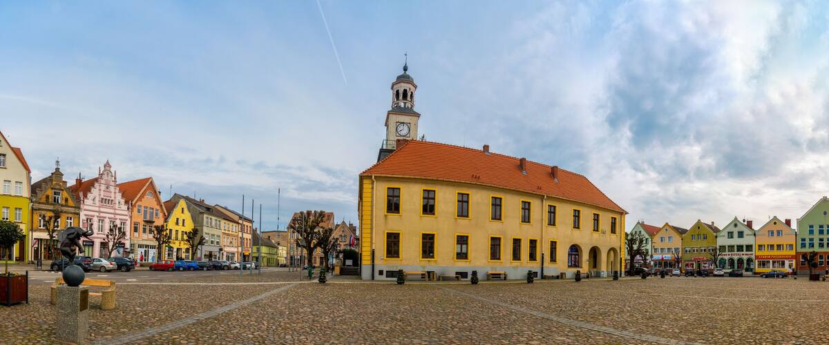 A cobblestone square with colorful buildings, a prominent clock tower, and a statue.