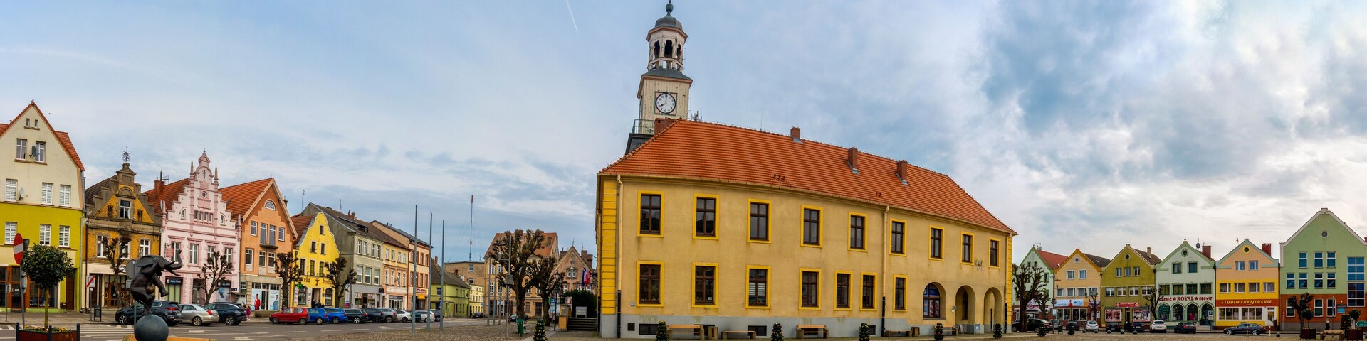A cobblestone square with colorful buildings, a prominent clock tower, and a statue.