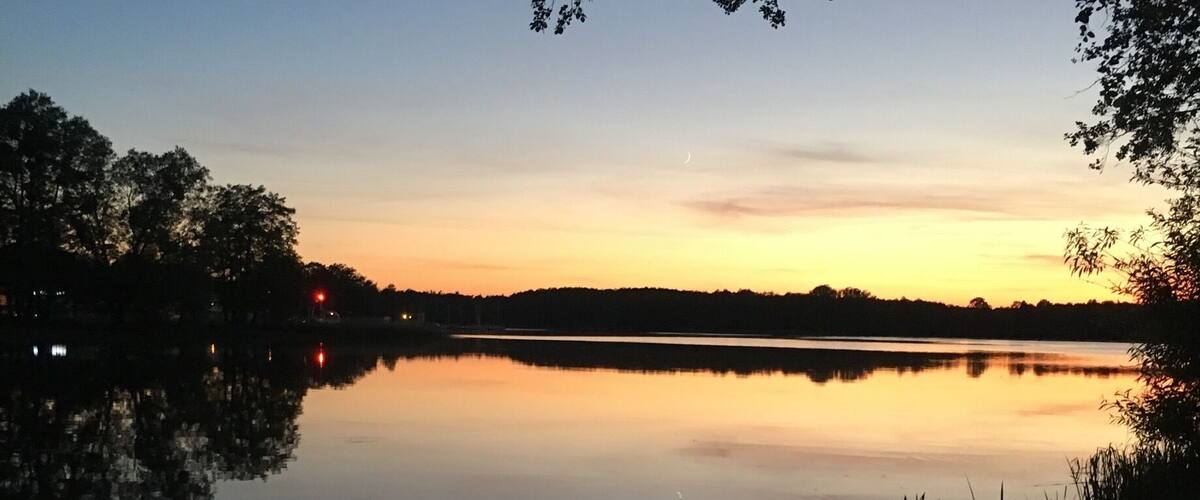 A calm lake at dusk with a clear sky, trees, and a visible crescent moon.