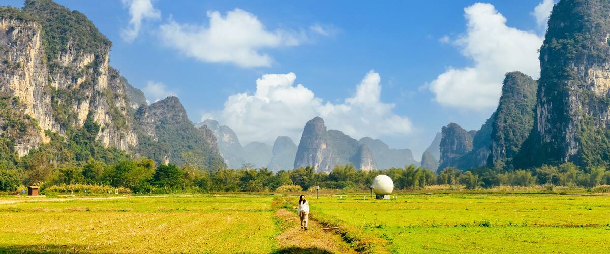 A person walking through a green field with a large spherical structure in the distance, surrounded by tall, rugged mountains.