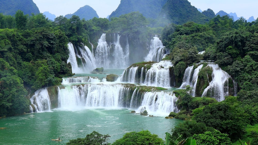 A waterfall cascading into a river surrounded by lush greenery and mountains.