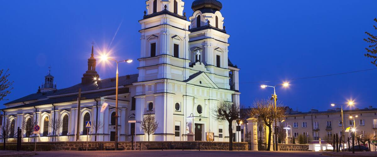 A church with two towers and a bell tower, surrounded by a paved area and street lamps.