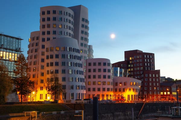 Eine Stadtlandschaft mit moderner Architektur, darunter Gebäude mit gebogenen und kantigen Formen, vor einem Dämmerhimmel.