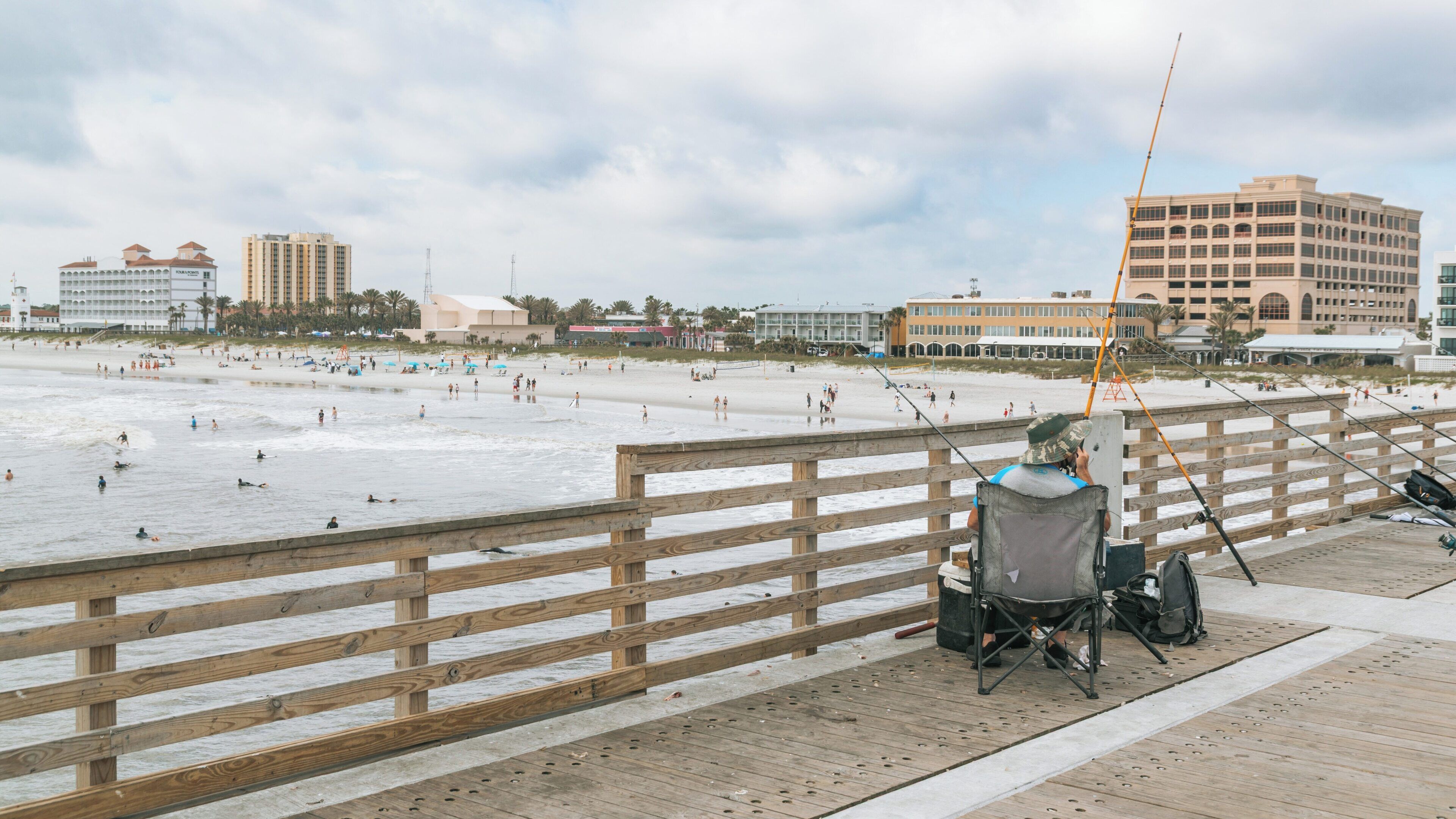 Fishing at Jacksonville Beach Pier on a cloudy day with surfers in the waves and people enjoying the sandy shore