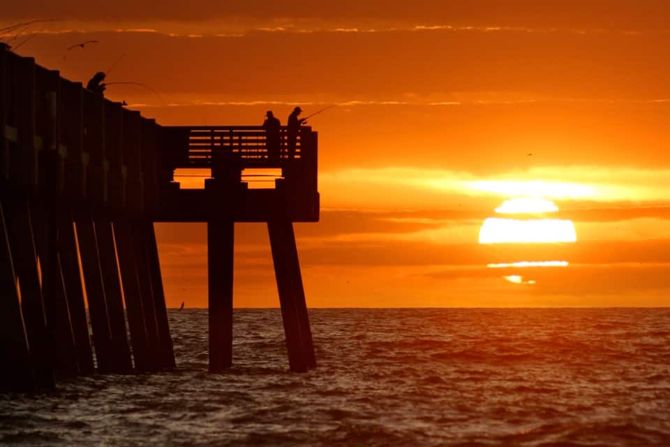 Um pôr do sol sobre o oceano, com um cais de madeira e duas pessoas pescando.